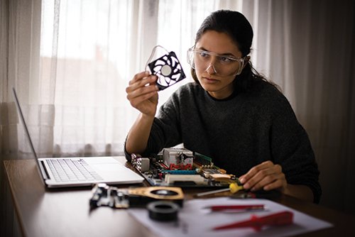 learning Photo of a student at home in front of a computer, wearing safety googles and examining a piece of computer hardware.