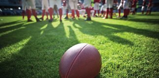 Photo of a football sitting on a field with players standing in the foreground