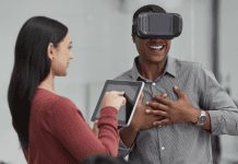 Photo of a man wearing a virtual reality headset while a woman holds an iPad next to him.