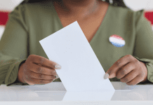 Photo of a Black woman pictured from neck down casting her vote in a ballot box while posed in front of American flag.